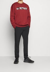 Uomo che indossa un felpa rossa Under Armour, pantaloni neri e sneakers nere Adidas, in piedi con le mani in tasca su uno sfondo bianco semplice.