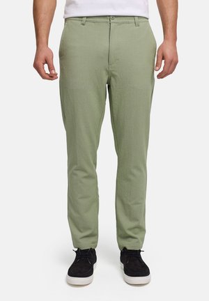 Hombre con pantalones texturizados de color verde claro, camisa blanca y zapatos casuales negros con cordones, de pie sobre un fondo claro y liso.