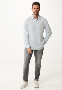 Světle modrá halenka s dlouhým rukávem a bez límce, nošená s šedými džíny a bílými teniskami. Materiál vypadá měkký a lehký.