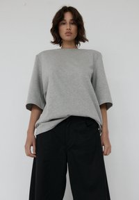 Jasnoszara, oversize koszulka z krótkim rękawem, wykonana z miękkiego materiału. Noszona z czarnymi szerokimi spodniami, obie na jednolitym tle.