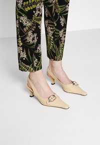 Escarpins slingback en cuir verni beige avec des pointes effilées, des talons blocs bas et des accents de boucle décoratifs sur le devant. Convient pour un usage décontracté ou formel.