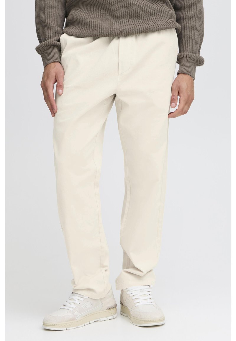 Pantalones chinos beige hechos de mezcla de algodón, con corte recto, bolsillos laterales y una textura sutil. Combinados con zapatillas blancas.