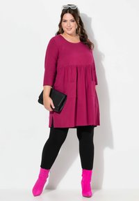 Vestido túnica magenta con cintura fruncida, dos bolsillos frontales, aberturas laterales, combinado con leggings negros y botas de tobillo rosas, sostenido con un clutch negro.