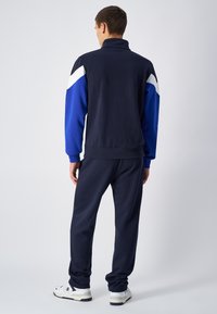 Tuta da ginnastica blu marina con accenti in blu e bianco. La giacca ha un colletto alto e maniche lunghe. I pantaloni sono tapered, realizzati in tessuto morbido, e presentano polsini elastici.