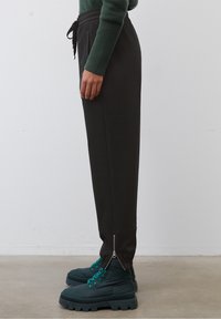 Joggers ajustados negros con detalles de cremallera en el bajo, combinados con botas de plataforma en tono verde azulado que llevan cordones en el mismo color, con un fondo neutro.