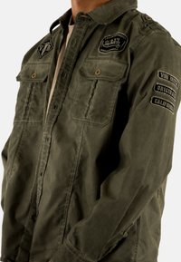 Giubbotto militare color verde oliva in cotone, dotato di due tasche sul petto, toppe ricamate e un design a vestibilità comoda.