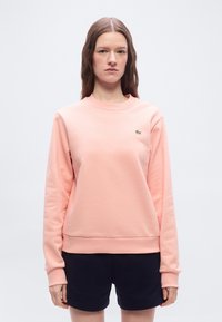 Γυναίκα με κυματιστά καστανά μαλλ ιά φοράει ροζ ανοιχτό, μακρυμάνικη μπλούζα και σκούρα μπλε σορτς, στέκεται μπροστά σε απλό λευκό φόντο.