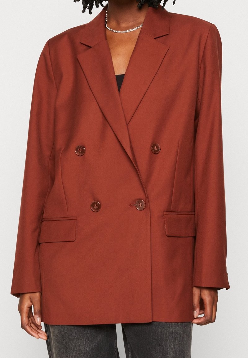 Femme portant un blazer croisé couleur rouille avec des revers en encoche et des poches à rabat, associé à un jean foncé et un collier chaîne argenté.