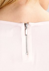 Světle růžová látka na zadní straně má kovový zip, který je částečně otevřený, s hladkým povrchem a malým taháčkem.