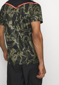 Hombre con una camisa de manga corta en camuflaje verde y negro, con ribete rojo en el hombro trasero, combinado con pantalones negros, visto desde atrás.