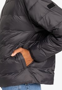Svart puffjacka med horisontell quiltad mönster, med en sidficka, elastiska ärmslut och strukturerad tyg. Diskret varumärkesetikett på ärmen.
