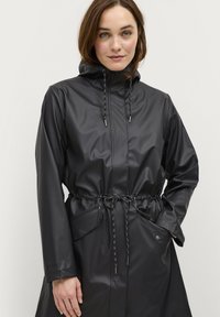 Cappotto impermeabile nero realizzato in materiale lucido, dotato di vita regolabile con coulisse, cappuccio e due tasche frontali con chiusura a pressione.