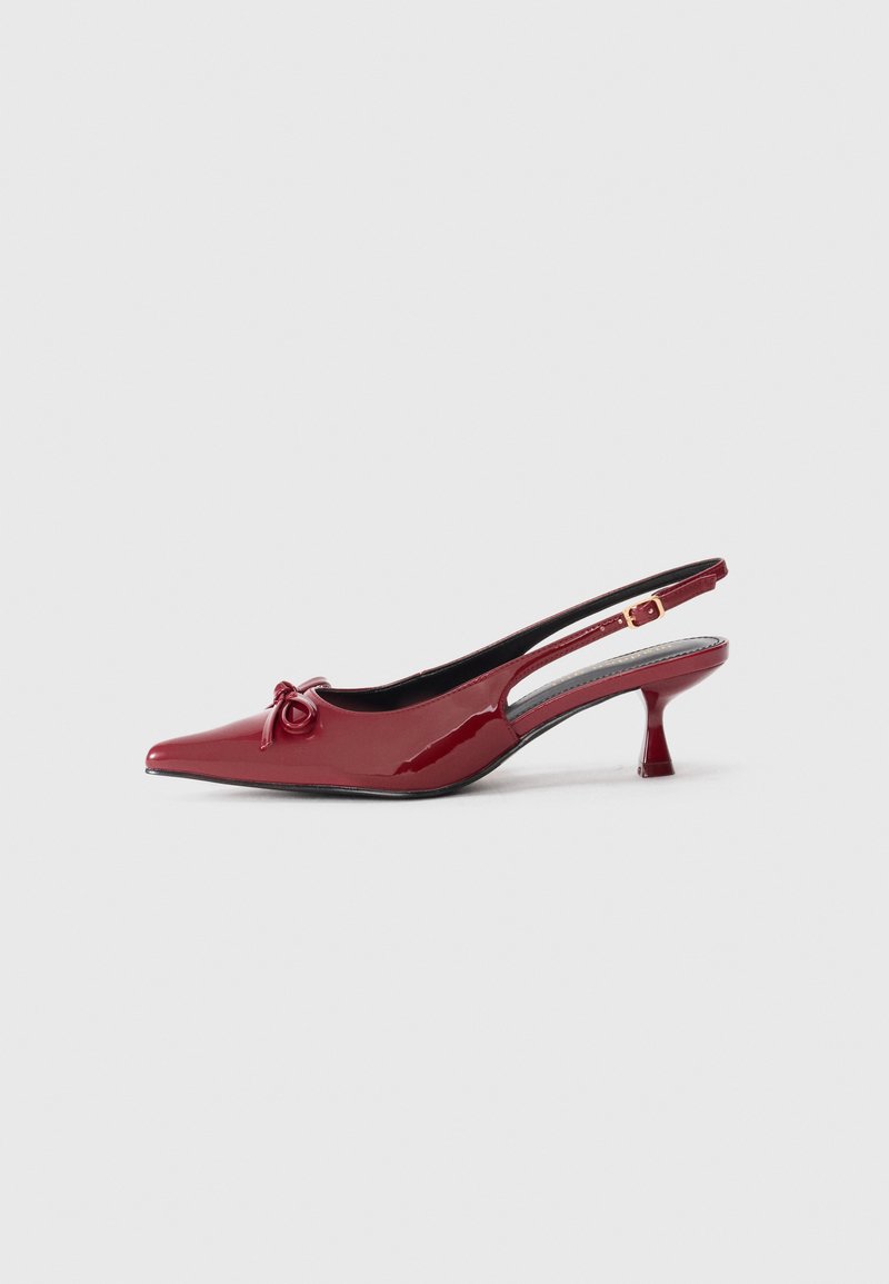 Sapatos slingback de verniz vermelho com biqueira pontiaguda, salto pequeno e um laço decorativo na parte da frente. Apresenta detalhe recortado nas laterais.