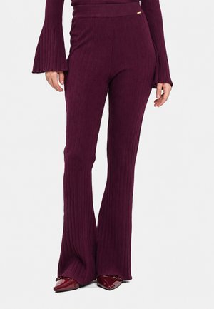 Donna che indossa pantaloni a vita alta a coste, a gamba larga, di colore viola scuro, abbinati a un top con maniche svasate coordinato e scarpe lucide con punta appuntita.