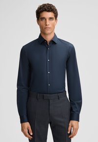 Navyblauwe shirt met lange mouwen, een knoopsluiting en een gladde textuur, gecombineerd met grijze broeken met een patroon.