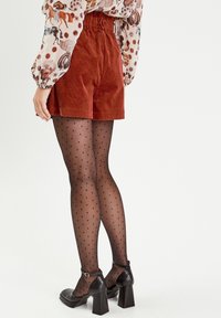 Rustfarbene Cordshorts mit hohem Bund, kombiniert mit transparenten schwarzen Strumpfhosen mit Punkten und schwarzen Schuhen mit Blockabsatz.