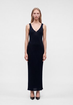 Femme debout portant une robe maxi noire côtelée sans manches et des talons noirs pointus sur un fond blanc uni.