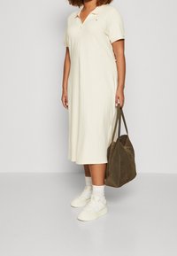 Vestido polo color crema hecho de una tela suave, con cuello y mangas cortas. Combinado con un bolso grande verde oscuro y zapatillas blancas.