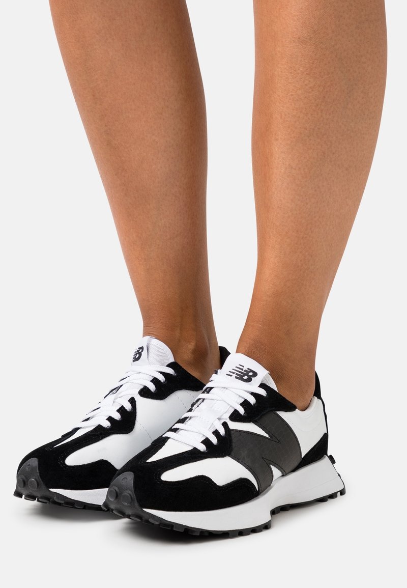 Jambes portant des baskets New Balance noires et blanches avec des semelles épaisses sur un fond clair uni.