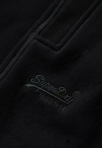 Zwarte fleece stof met een tonale geborduurde logo "SuperDry" en accentdetails. De textuur lijkt zacht en glad.