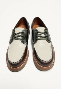 Barna, krémszínű és fekete bőrből készült hajócipő pár, spiccvarrással és zöld fűzőkkel, fehér háttéren felülről ábrázolva.