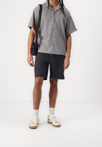 Man som bär en grå kortärmad skjorta med knappar, svarta denimshorts, vita sneakers och bär en svart axelväska.