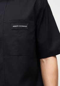 Chemise noire à manches courtes avec l'étiquette "ARMANI EXCHANGE" cousue au-dessus de la poche poitrine, portée par une personne dont le bras est visible sous la manche.