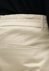 Pantalon beige avec une taille élastique et une poche passepoilée arrière, porté avec un pull en maille noir.