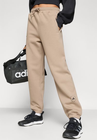 Μπεζ φόρμες με ελαστική μέση και μανσέτες, με πλαϊνές τσέπες και κυκλικό λογότυπο στον αστράγαλο. Μαύρα αθλητικά παπούτσια και τσάντα.