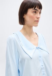 Γυναίκα με κοντά σκούρα μαλλιά, φορώντας ανοιχτό μπλε μπλούζα με φαρδύ γιακά και διακοσμητική ασημένια λεπτομέρεια, στραμμένη προς τα δεξιά σε απλό φόντο.