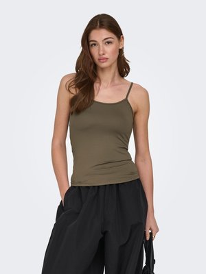 Young woman with long brown hair wearing an olive green spaghetti strap top and loose black pants, holding a black bag in her right hand.