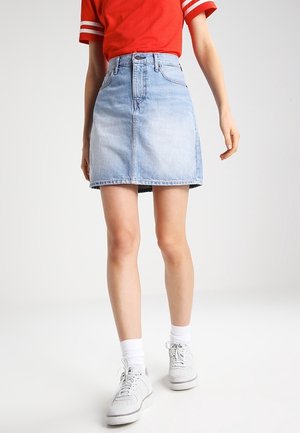 Personne portant un t-shirt rouge à manches courtes, une jupe en denim bleu clair, des chaussettes blanches et des baskets blanches, debout sur un fond clair uni.