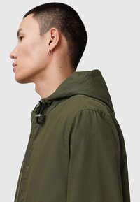 Veste à capuche vert olive en matière légère. Dispose d'une fermeture éclair à l'avant, d'une capuche ajustable et d'un design minimaliste.
