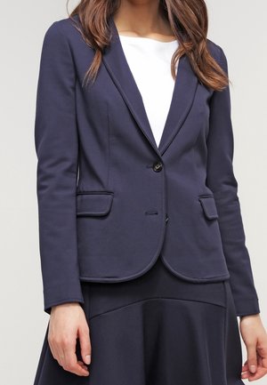 Vrouw draagt een marineblauw aansluitend blazer met klepzakken en een wit topje eronder, zichtbaar van schouders tot halverwege de dijen.