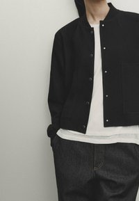 Personne portant une veste noire boutonnée sur une chemise blanche, avec un jean foncé et les mains dans les poches, devant un mur clair uni.