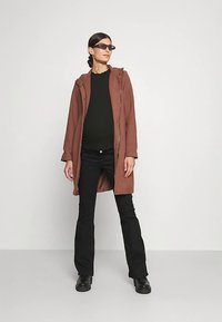 Hosszú barna kabát kapucnival, fekete felső és fekete, bő szárú nadrág fölött viselve. Fekete csizmát és téglalap alakú napszemüveget visel. Egyszínű háttér.