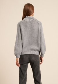 Femme aux longs cheveux bruns portant un gros pull tricoté gris clair et un pantalon en velours côtelé gris foncé, debout de dos sur un fond beige.