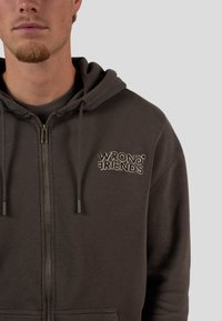 Tmavě šedá mikina na zip s kapsou na přední straně a vyšívaným textem "WRONG FRIENDS" v kontrastní béžové barvě na levé hrudi.