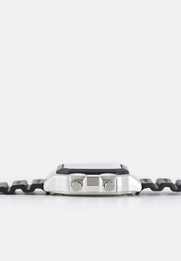 Montre numérique avec un boîtier en métal argenté, des accents en caoutchouc noir et un bracelet noir de forme irrégulière. Dispose de deux boutons latéraux.