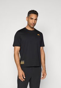 Svart t-skjorte med korte ermer laget av mykt stoff, med en oransje logo på brystet og et mønstrete aksent på kanten.