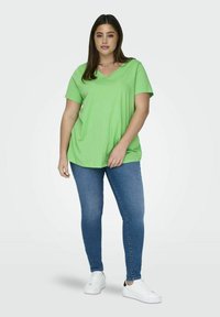 Zöld, rövid ujjú, V nyakú póló, laza fazonban, kék, skinny farmernadrággal és fehér sportcipővel egy semleges háttéren.
