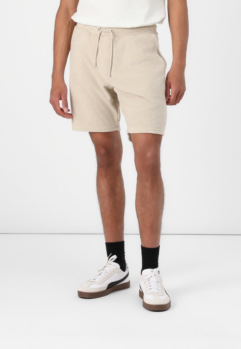 Pantalones cortos de toalla beige con cintura elástica, cierre con cordón y bolsillos laterales. Combinados con zapatillas blancas y calcetines negros.