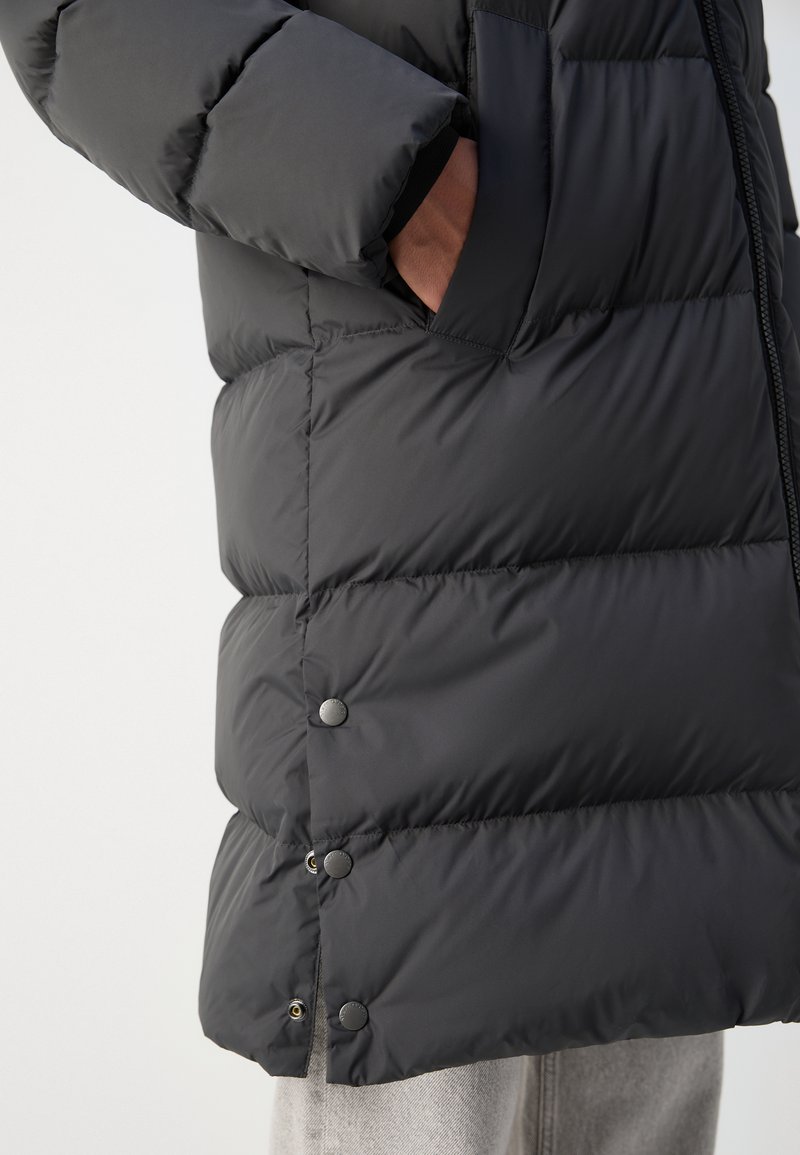 Chaqueta de plumas negra con un diseño acolchado, presenta botones a presión metálicos y un bolsillo lateral. Tejido suave y resistente al agua.