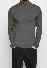 Gray long-sleeve turtleneck shirt made of soft fabric, fitted design, and a smooth texture. Worn with black pants, hands in pockets.
