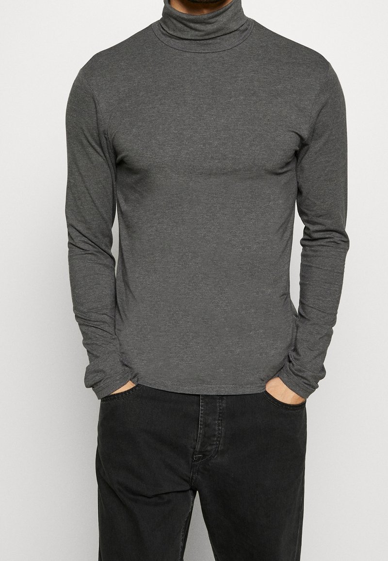 Gray long-sleeve turtleneck shirt made of soft fabric, fitted design, and a smooth texture. Worn with black pants, hands in pockets.