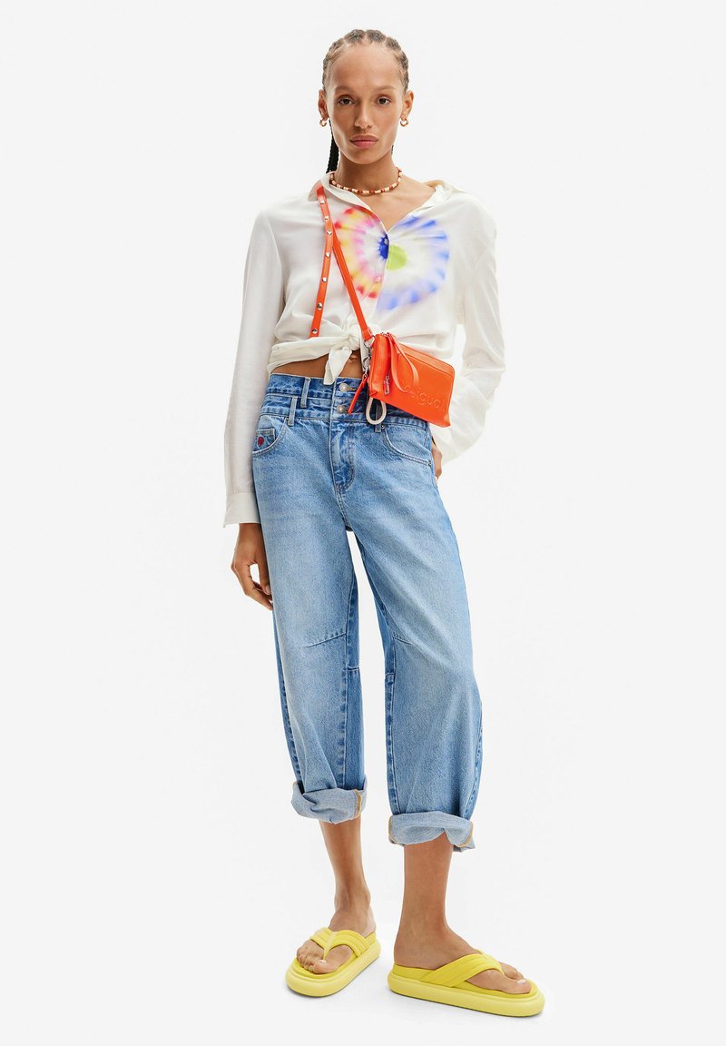 Camicia bianca a maniche lunghe con un motivo tie-dye, jeans oversize blu chiaro e ciabatte gialle. Borsa arancione a tracolla con marchio.