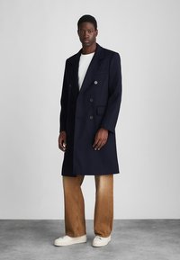 Cappotto doppiopetto navy in tessuto testurizzato, caratterizzato da grandi bottoni e colletto a revers, abbinato a pantaloni beige e scarpe da ginnastica bianche.