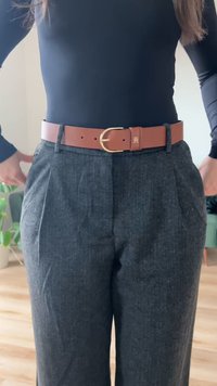Pantaloni grigi a spina di pesce con pieghe laterali, abbinati a una cintura in pelle marrone con fibbia dorata. Lo sfondo presenta della vegetazione.
