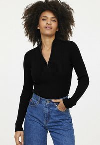 Schwarzer Ripp-Pullover mit Halbzipp-Kragen, taillierte Passform. Das Modell trägt hochtaillierte Jeans, was einen lässigen Stil unterstreicht.