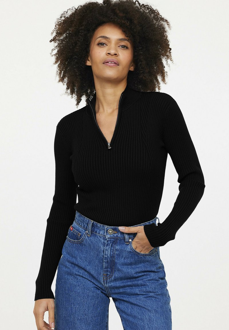 Schwarzer Ripp-Pullover mit Halbzipp-Kragen, taillierte Passform. Das Modell trägt hochtaillierte Jeans, was einen lässigen Stil unterstreicht.